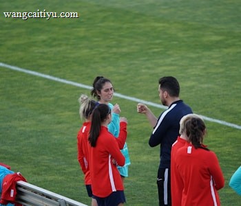 A group of people wearing sports attire are gathered on a grassy field. Some individuals are dressed in red jackets, while others are in turquoise and navy blue. A coach appears to be giving instructions. Sports equipment, such as water bottles and a bench with towels, can be seen nearby.