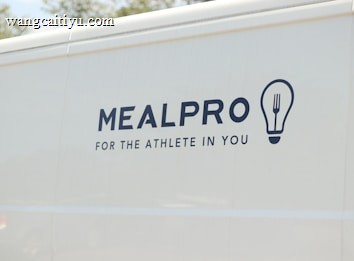 The side of a white vehicle displays the meal service logo 'MEALPRO' with the tagline 'For the Athlete in You'. Next to the text is a graphic combining a lightbulb and a fork.